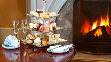 A three-tiered dessert stand with various pastries and sandwiches, a tea set, and glasses of water on a table with a fireplace in the background.