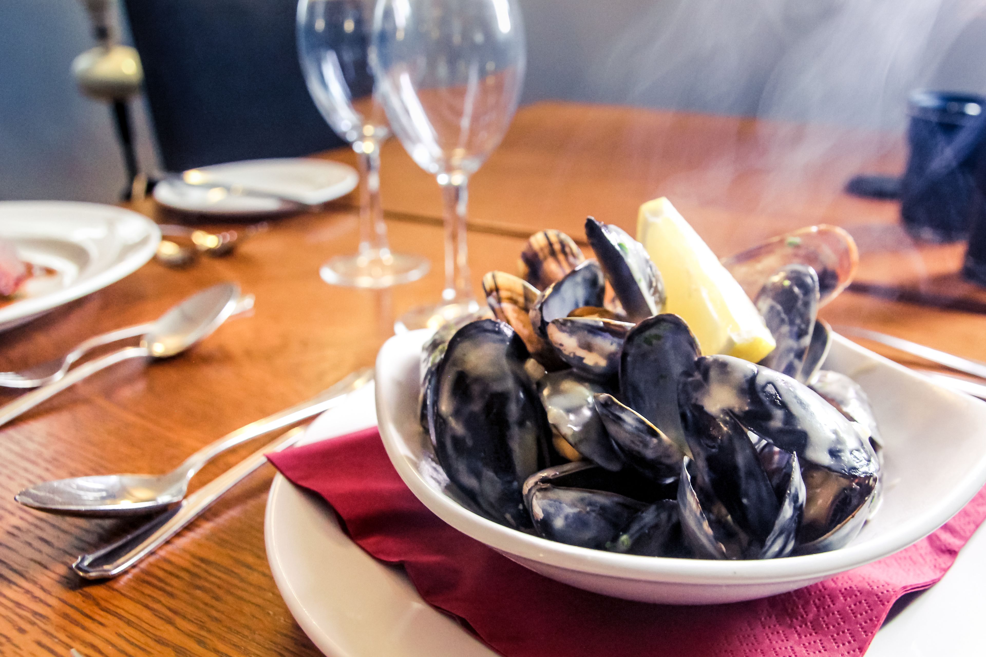 A bowl of mussels on a table with a red napkin, wine glasses, and cutlery.