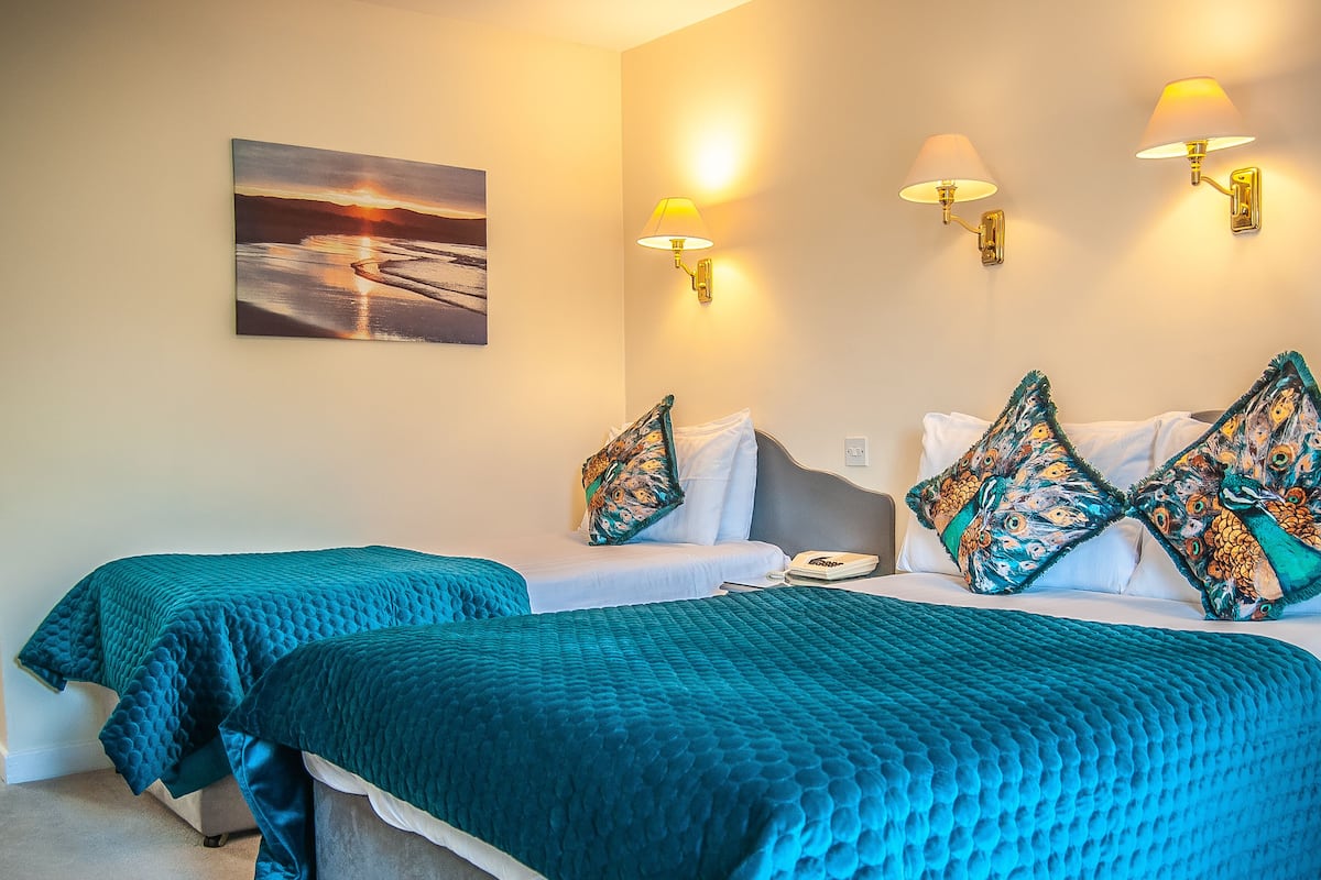 Une chambre d’hôtel avec deux lits, une literie de couleur turquoise, un tableau représentant un coucher de soleil et des lampes fixées au mur.