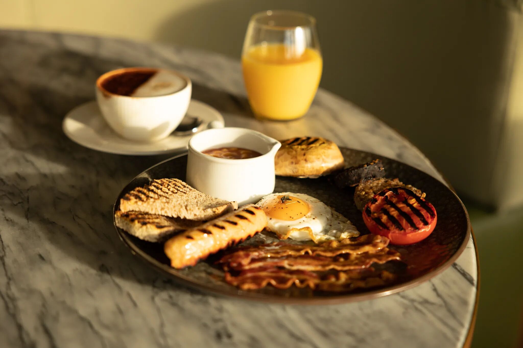 En morgenmadstallerken med ristet toast, bacon, et spejlæg og en grillet tomat på et marmorbord.