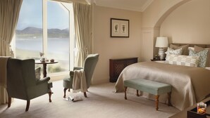 A bedroom with a large bed, a sitting area with chairs, a table with a vase, and a view of the sea.