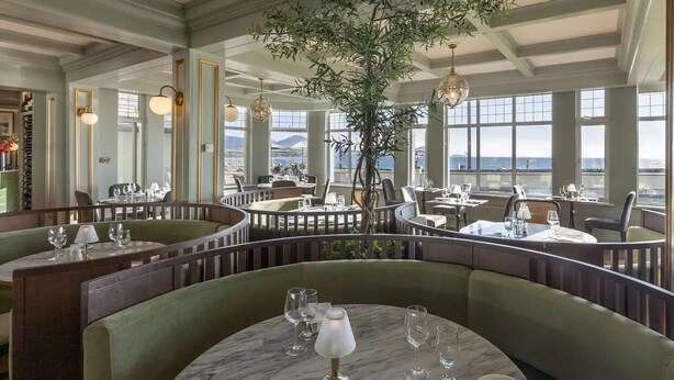 A restaurant with round tables and booths, large windows offering a view of the sea, and a hanging plant in the center.