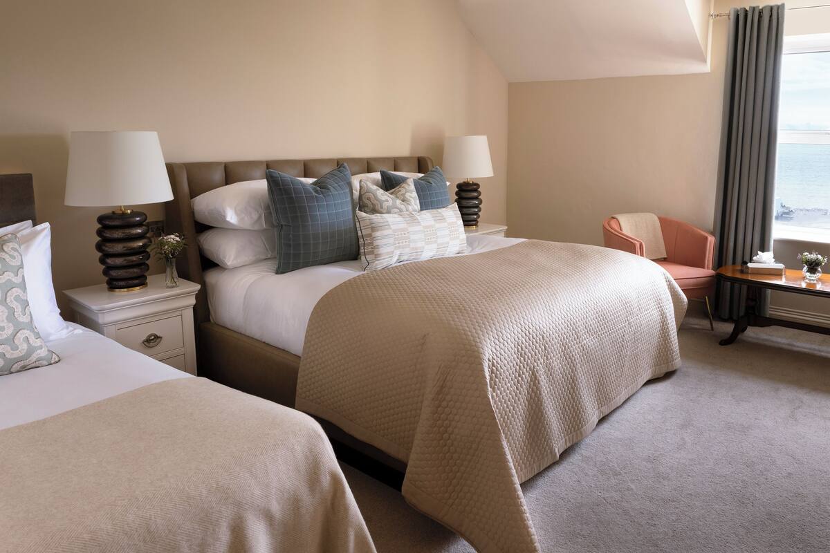 A hotel room with two beds, a view of the sea, a chair, and a table with a lamp.