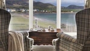 A room with a view of a coastal landscape, featuring two plaid armchairs, a wooden side table with a bottle, a glass, and a teapot.