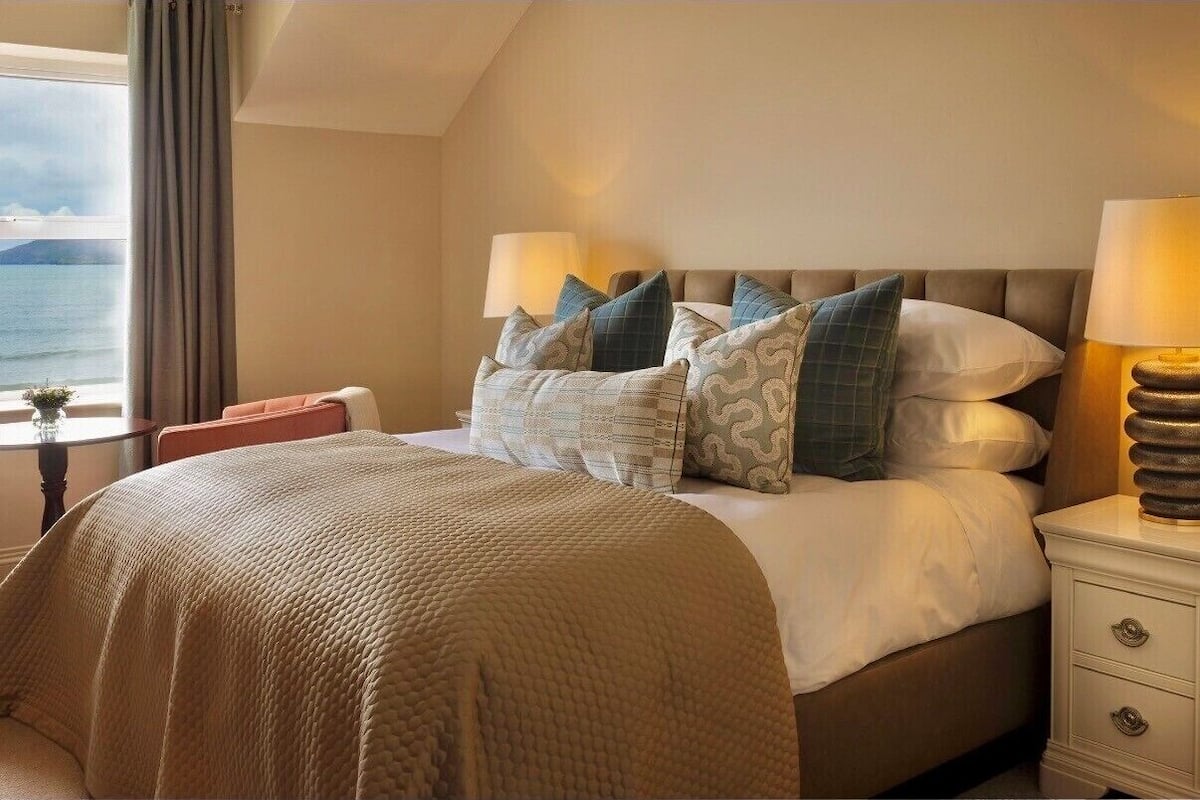 A hotel room with a bed, two bedside lamps, a nightstand, a window with a view of the sea, and a small table with a vase.