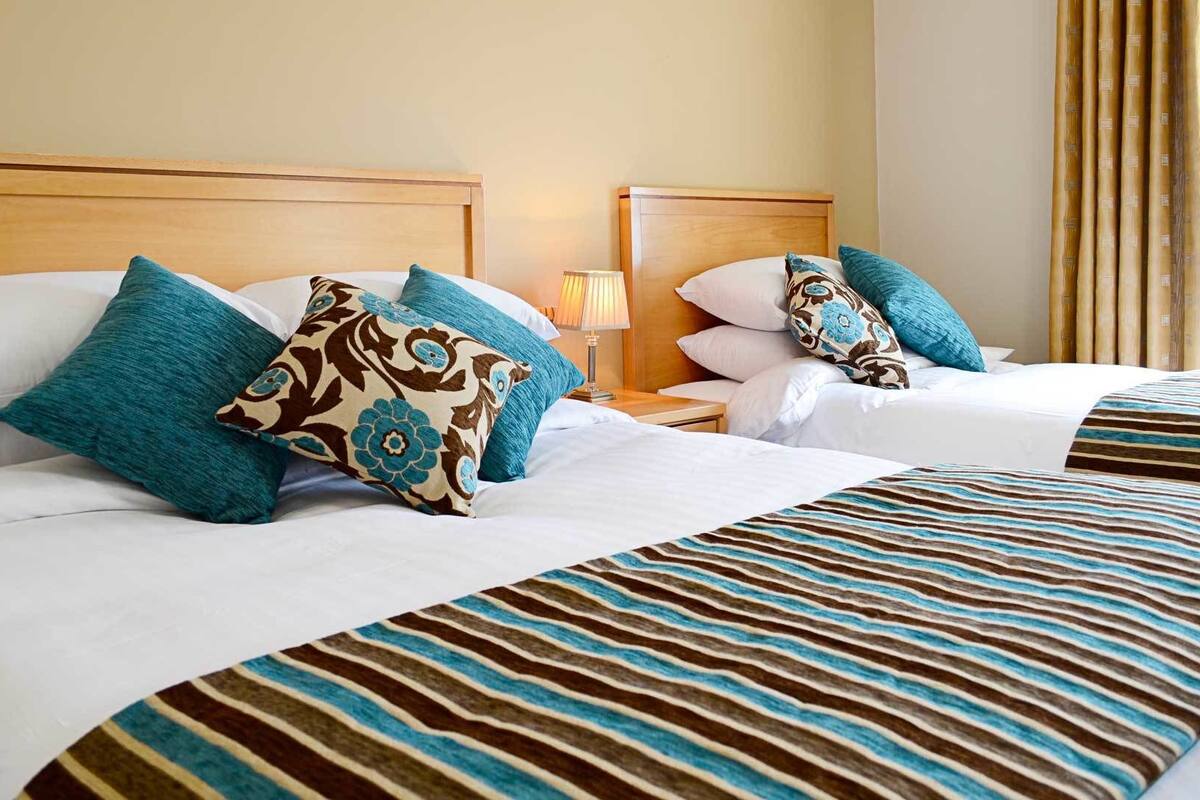 A hotel room with two beds, each with white linens and striped bedspreads. There are multiple pillows with floral patterns and solid colors. A bedside table with a lamp is visible.