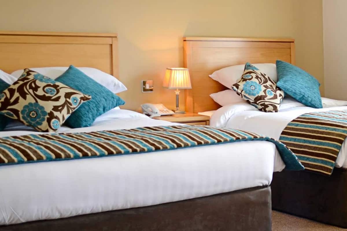 A hotel room with two beds, each with white bedding and teal pillows. There is a bedside table with a lamp and a phone.