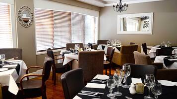 A dining area with tables set for multiple guests, large windows with blinds, and a chandelier.
