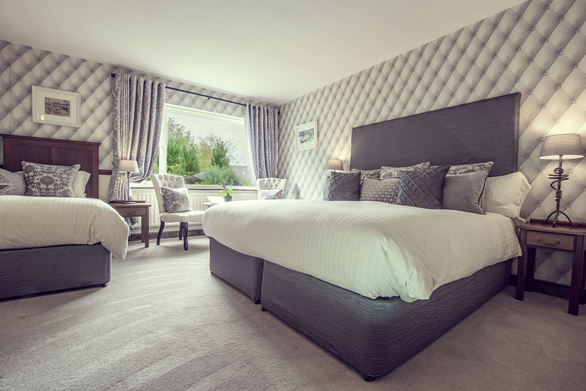 A hotel room with two beds, a sitting area with a chair, a window with curtains, and a view of greenery outside.
