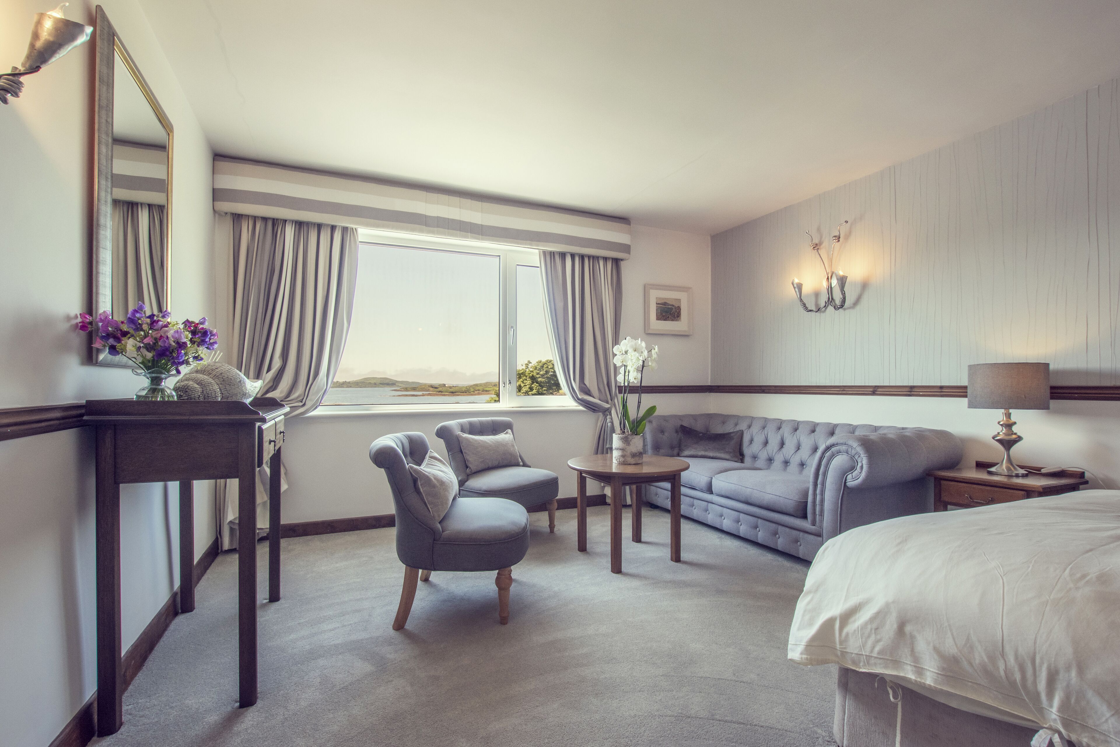 Habitación de hotel con una cama grande, un sofá, dos sillones, una mesita con un jarrón de flores, una lámpara y un ventanal con cortinas.