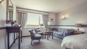 Habitación de hotel con una cama grande, un sofá, dos sillones, una mesita con un jarrón de flores, una lámpara y un ventanal con cortinas.