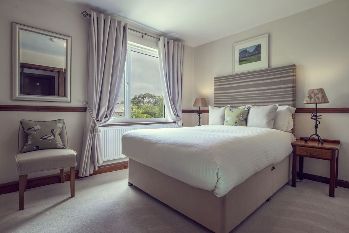 A bedroom with a large bed, a chair, a nightstand, a lamp, a window with curtains, and a framed picture on the wall.