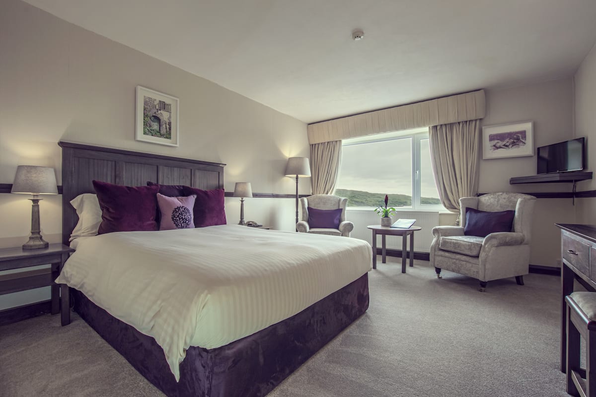 A spacious hotel room with a large bed, two armchairs, a small table, a TV, and a window with curtains.