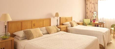 A hotel room with two beds, wooden headboards, bedside tables with lamps, a small table with a vase of flowers, and a chair.