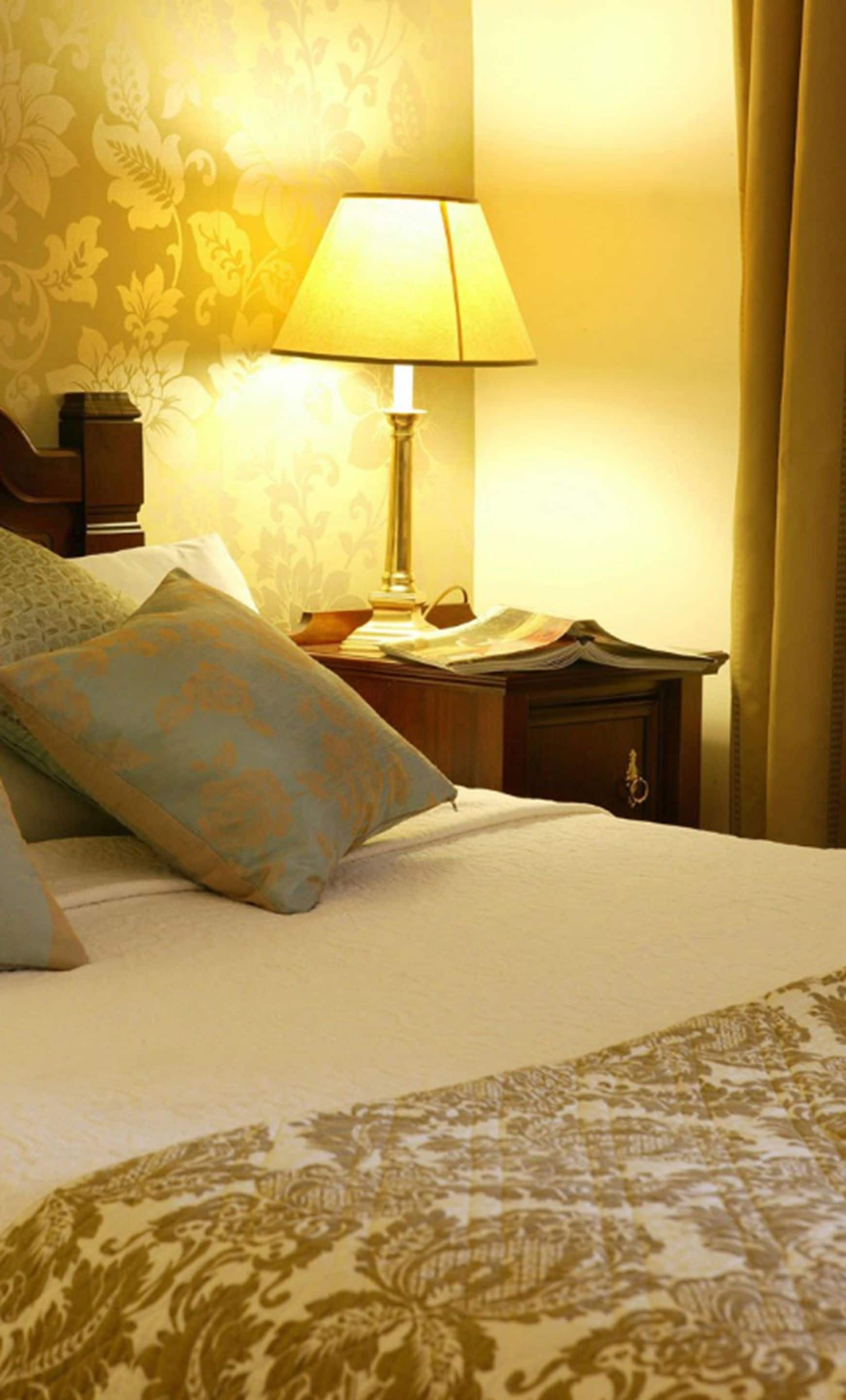 A neatly made bed with a patterned bedspread, a bedside lamp, and a nightstand with a book and papers.