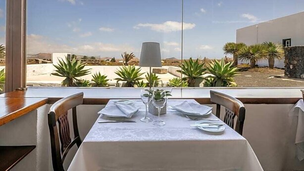 A dining table set with white tablecloth, plates, and cutlery, with a lamp and potted plants on the table.