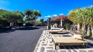 A stone pathway leading to a seating area with lounge chairs and tables, surrounded by tropical plants and a clear blue sky.