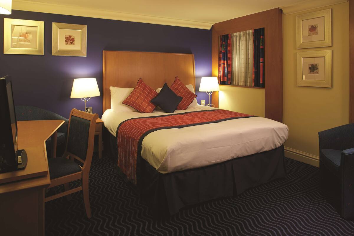 A hotel room with a large bed, two bedside lamps, a desk with a chair, and a television.