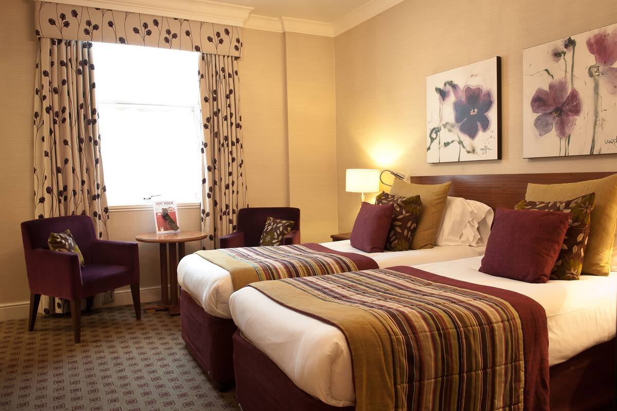 A hotel room with two beds, a chair, a small table, and floral artwork on the wall.