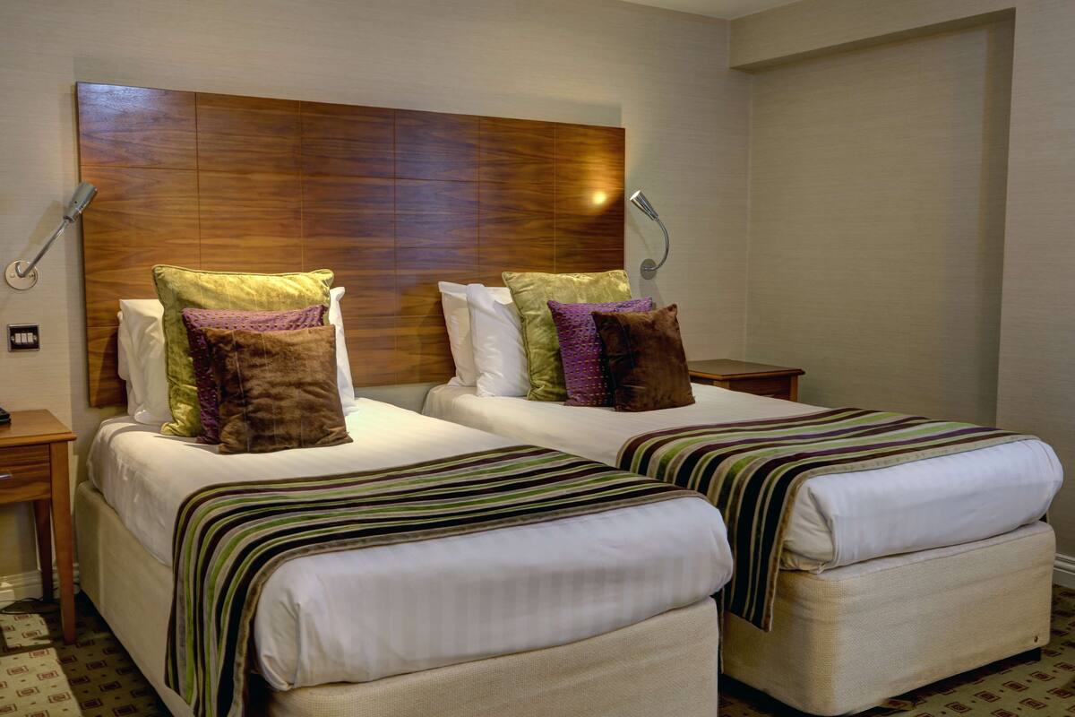 A hotel room with two beds, wooden headboard, bedside lamps, and a patterned rug.