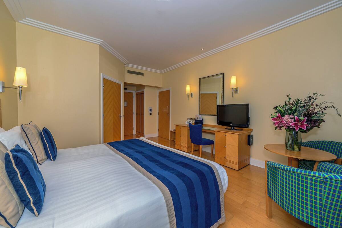 A hotel room with a large bed, a desk with a chair, a television, and a small table with a vase of flowers.