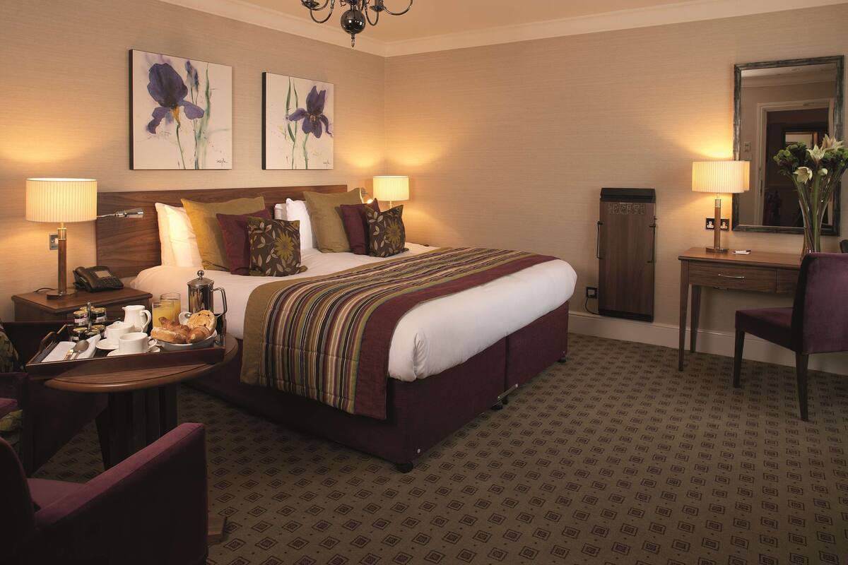 A hotel room with a bed, bedside lamps, a desk with a chair, a small table with breakfast items, and artwork on the walls.