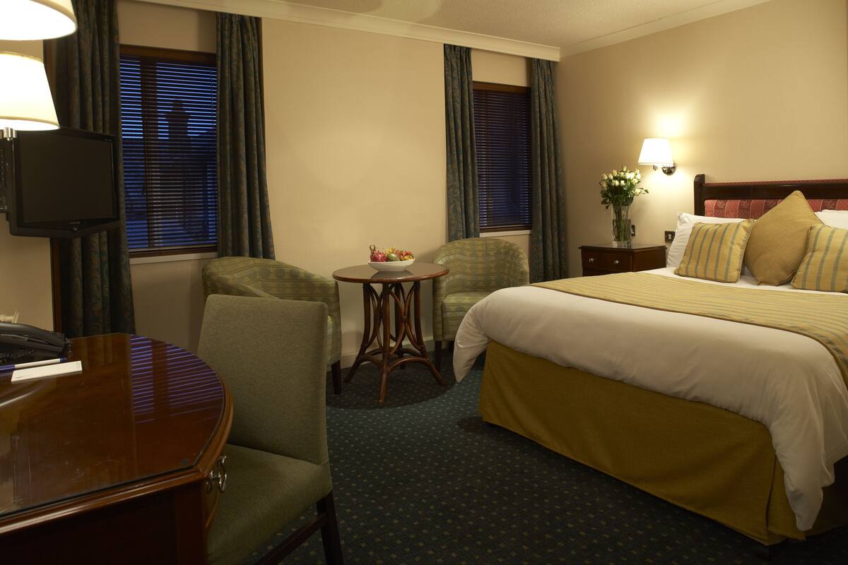 A hotel room with a bed, two armchairs, a small table, a television, and a nightstand with a lamp.