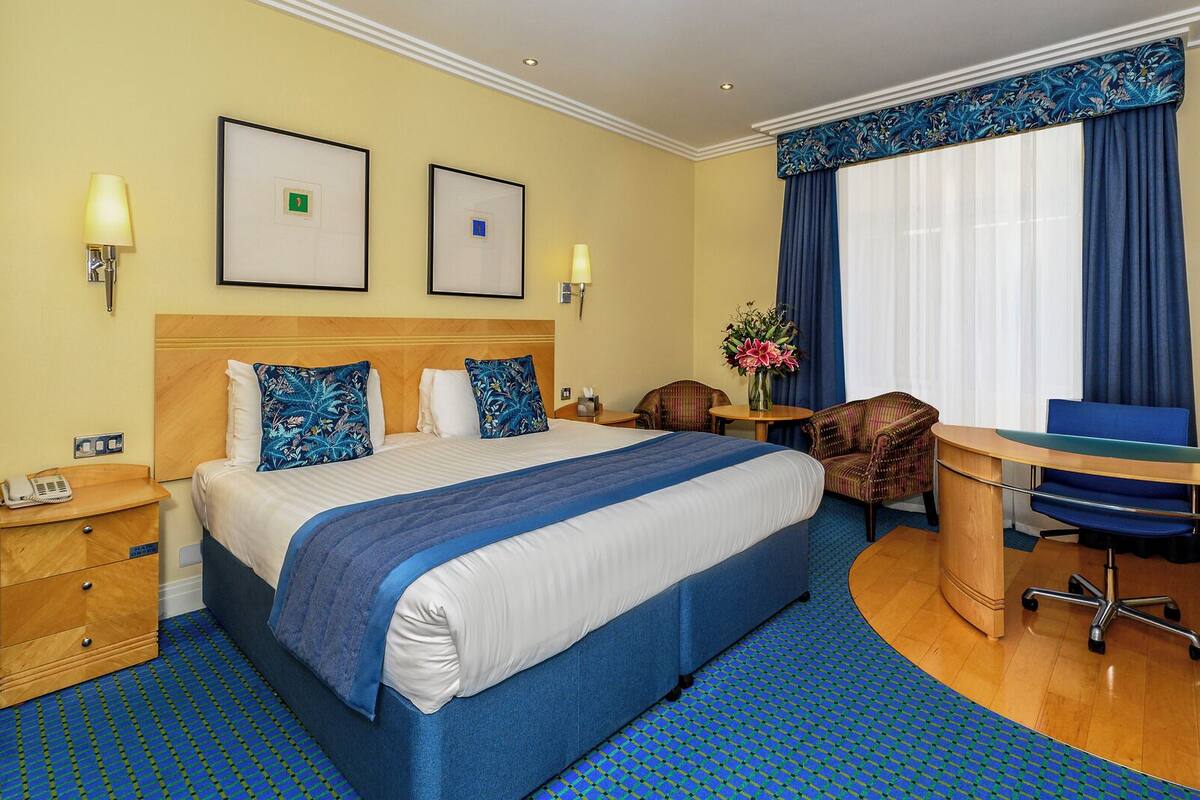 A hotel room with a large bed, two bedside tables, a desk, a chair, and a large window with curtains.