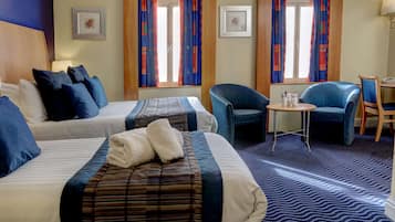 A hotel room with two beds, a small table, two chairs, and a view of the outside through patterned curtains.
