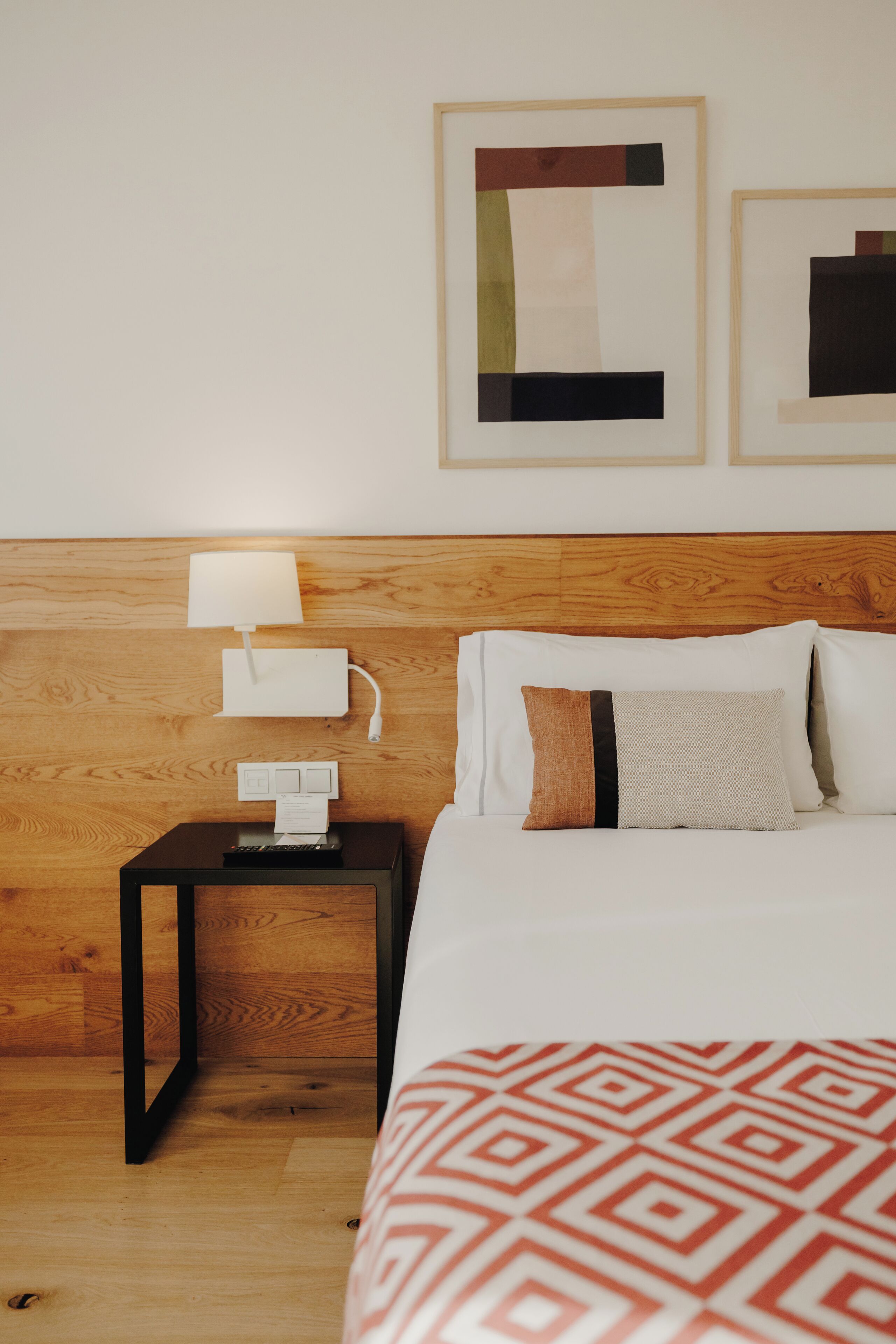 A hotel room with a wooden headboard, a bedside table with a lamp and phone, a bed with patterned bedding, and two framed abstract artworks on the wall.