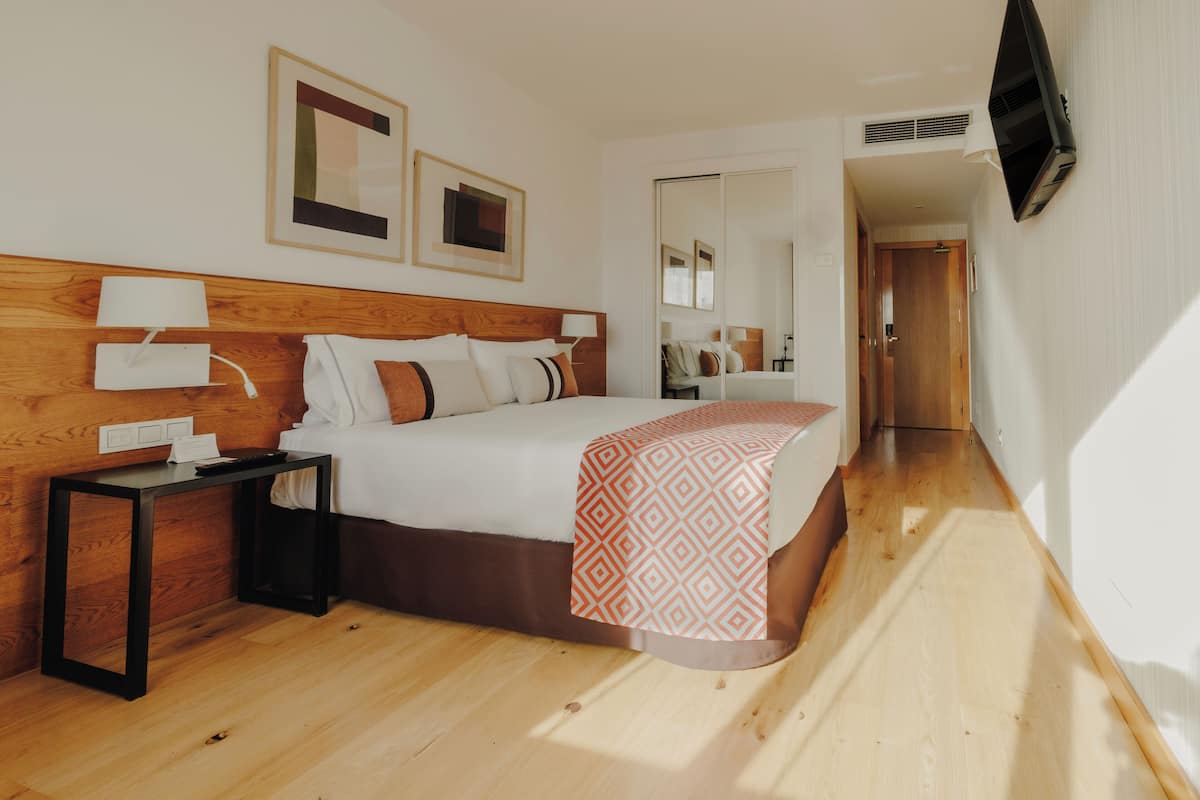 A hotel room with a wooden headboard, a bed with a patterned blanket, a nightstand with a lamp, and a flat-screen TV mounted on the wall.