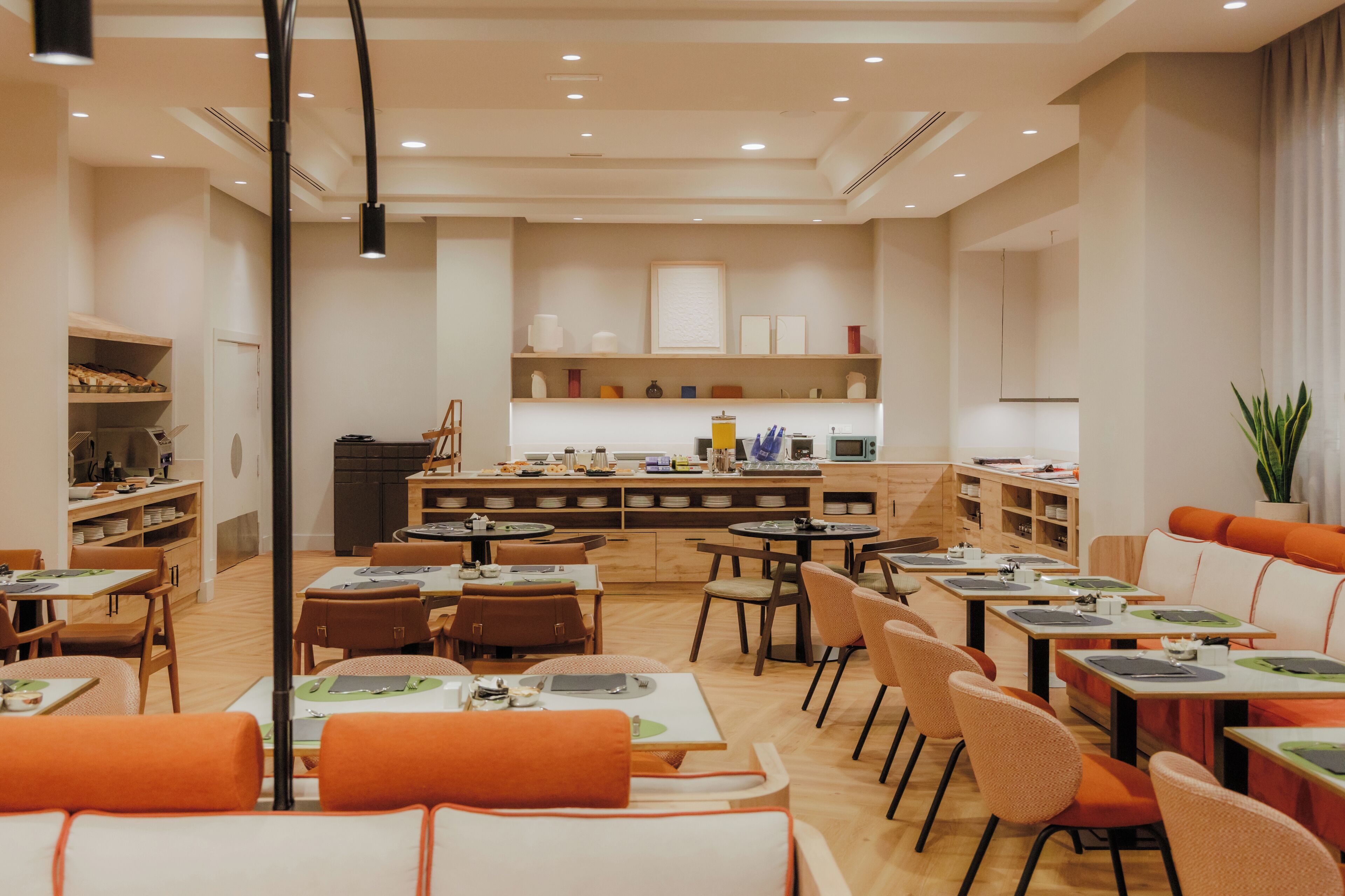 A modern dining area with multiple tables and chairs, a kitchen in the background, and shelves with various items.