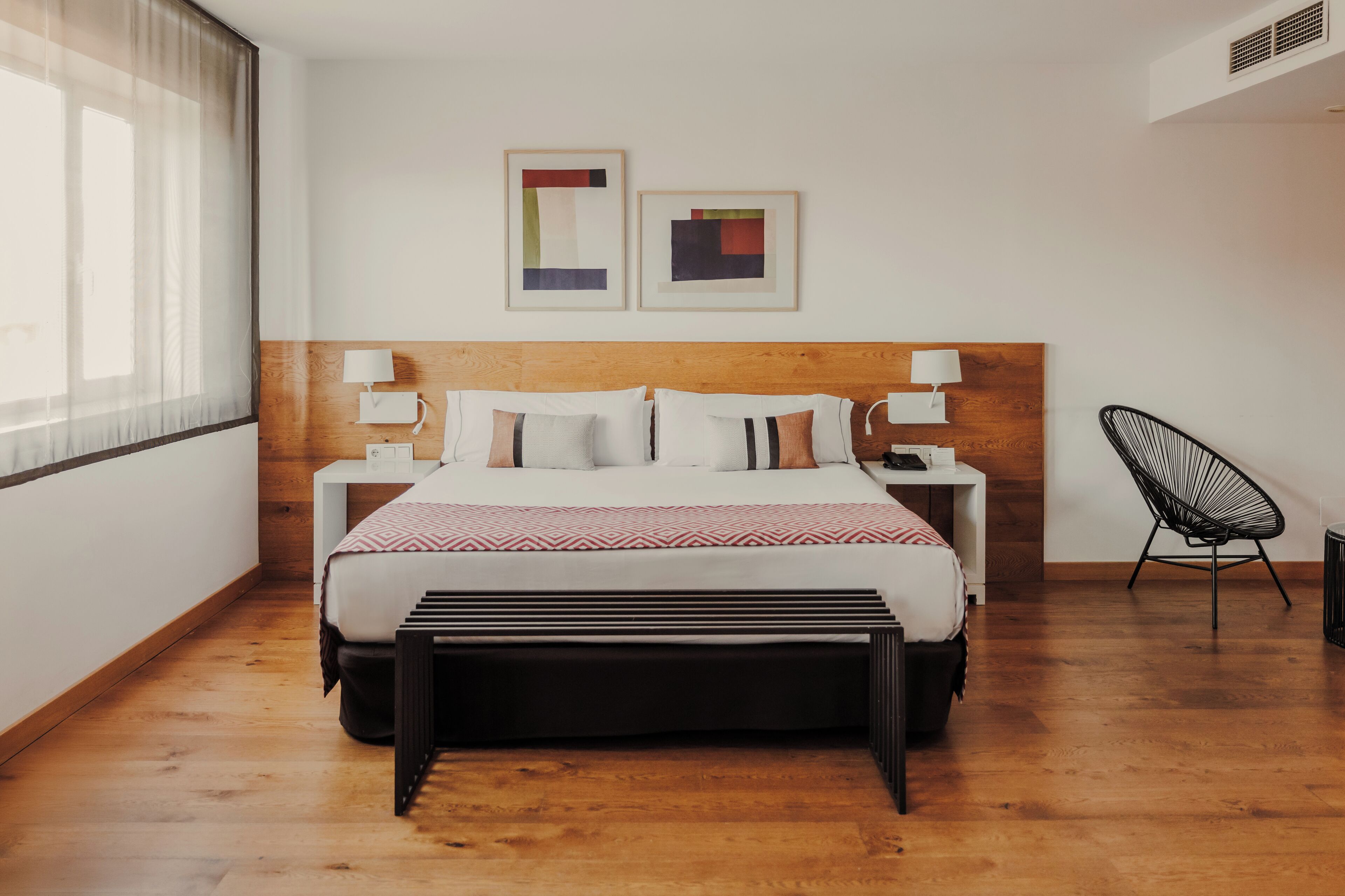 A modern hotel room with a wooden headboard, a large bed with white bedding, a bedside table with a lamp, a black chair, and two abstract paintings on the wall.