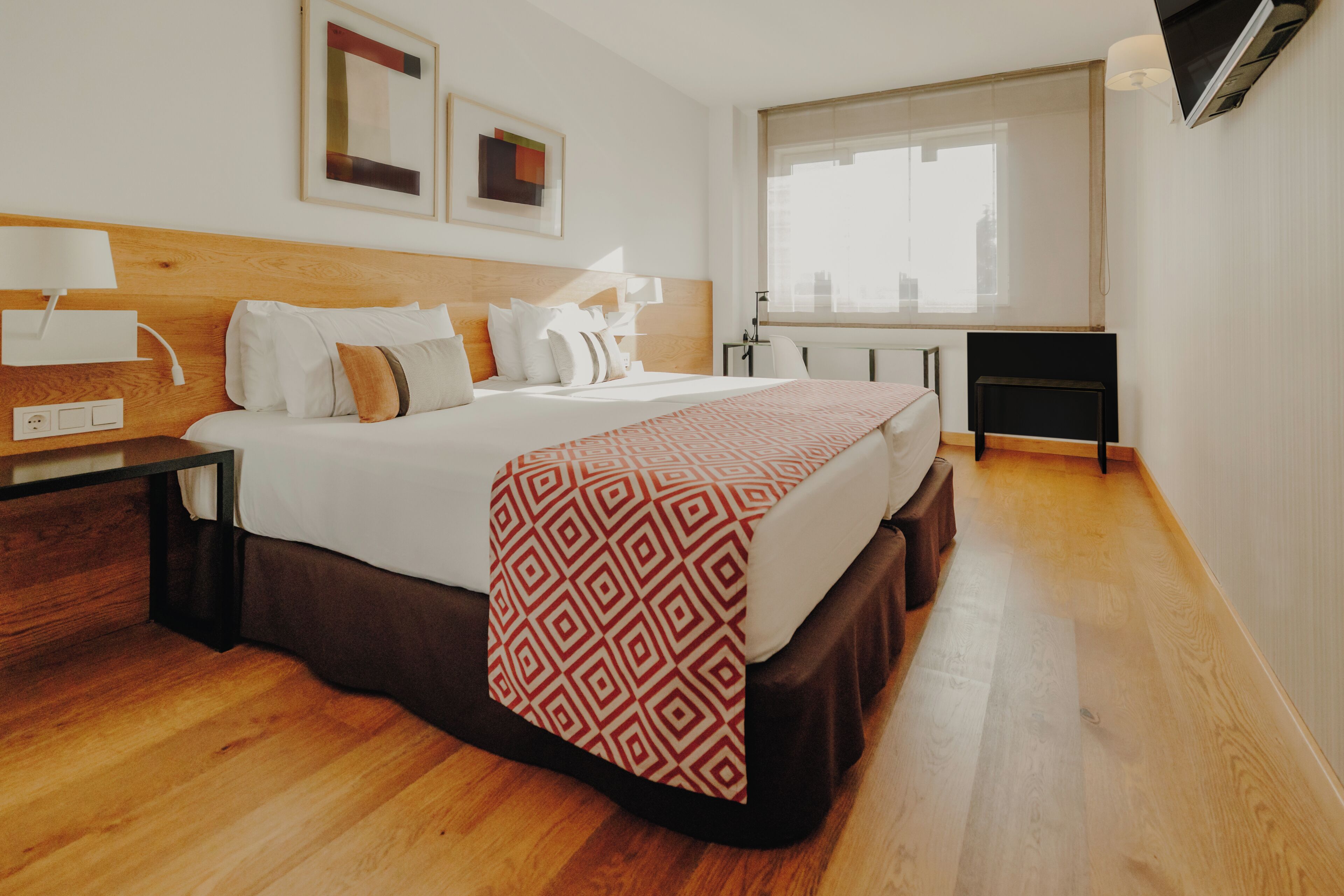 Habitación de hotel con cabecera de madera, cama grande con ropa de cama estampada, mesita de noche con lámpara, escritorio con silla y ventana con vistas a edificios.