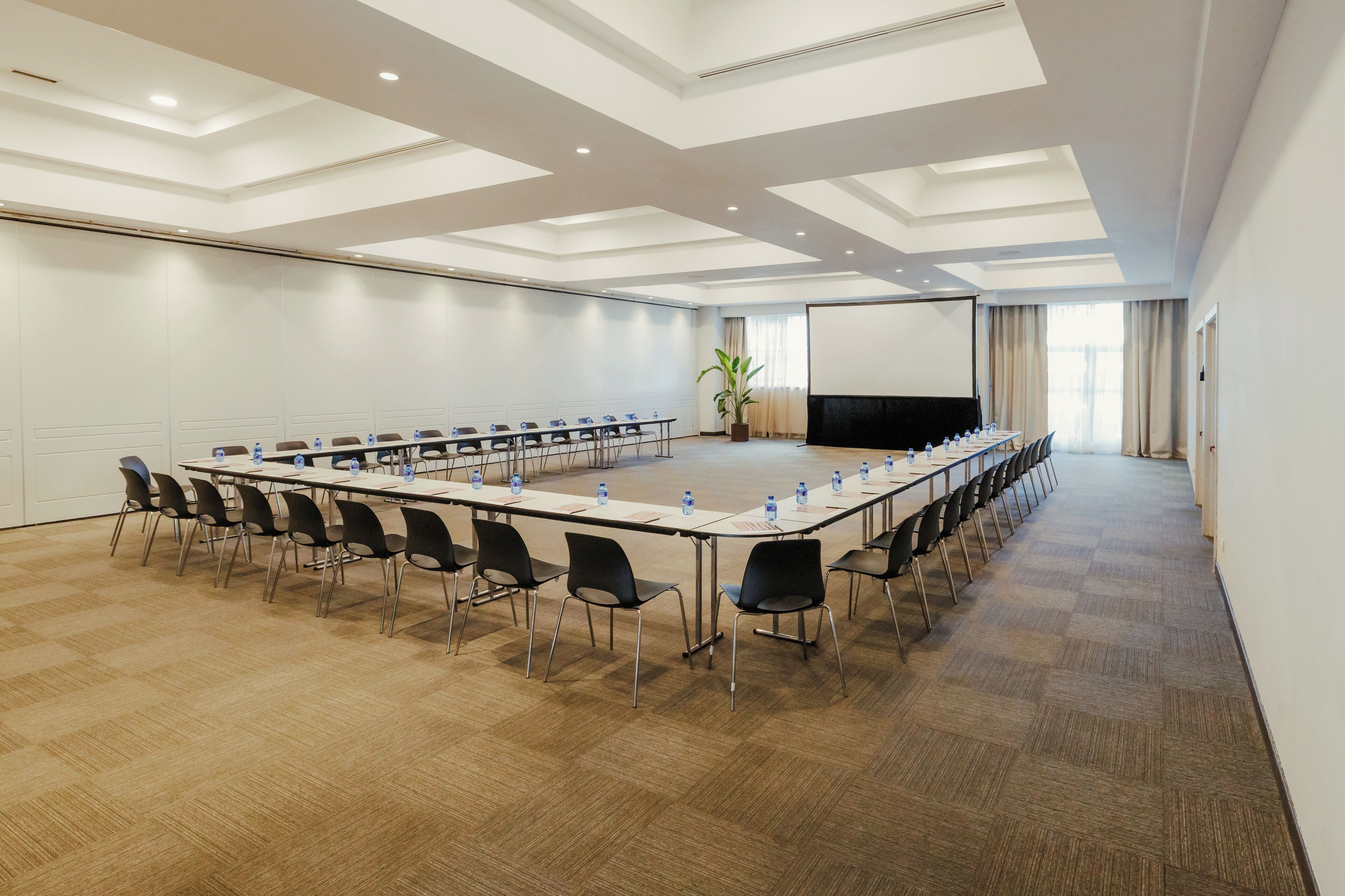 Un salón de conferencias con una larga mesa, sillas, una pantalla de proyector y una planta grande.