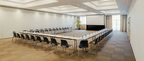 A conference room with a long table, chairs, a projector screen, and a large plant.