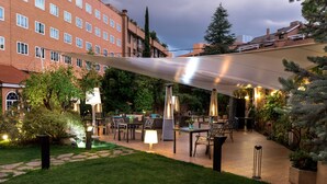 Un espace extérieur aménagé en terrasse, avec des tables et des chaises, entouré de verdure et doté d’un grand parasol pour créer de l’ombre.