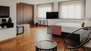 Une chambre d’hôtel moderne avec un plancher en bois, un lit recouvert d’un couvre-lit à motifs, une chaise noire, une petite table, un téléviseur et un téléviseur fixé au mur.