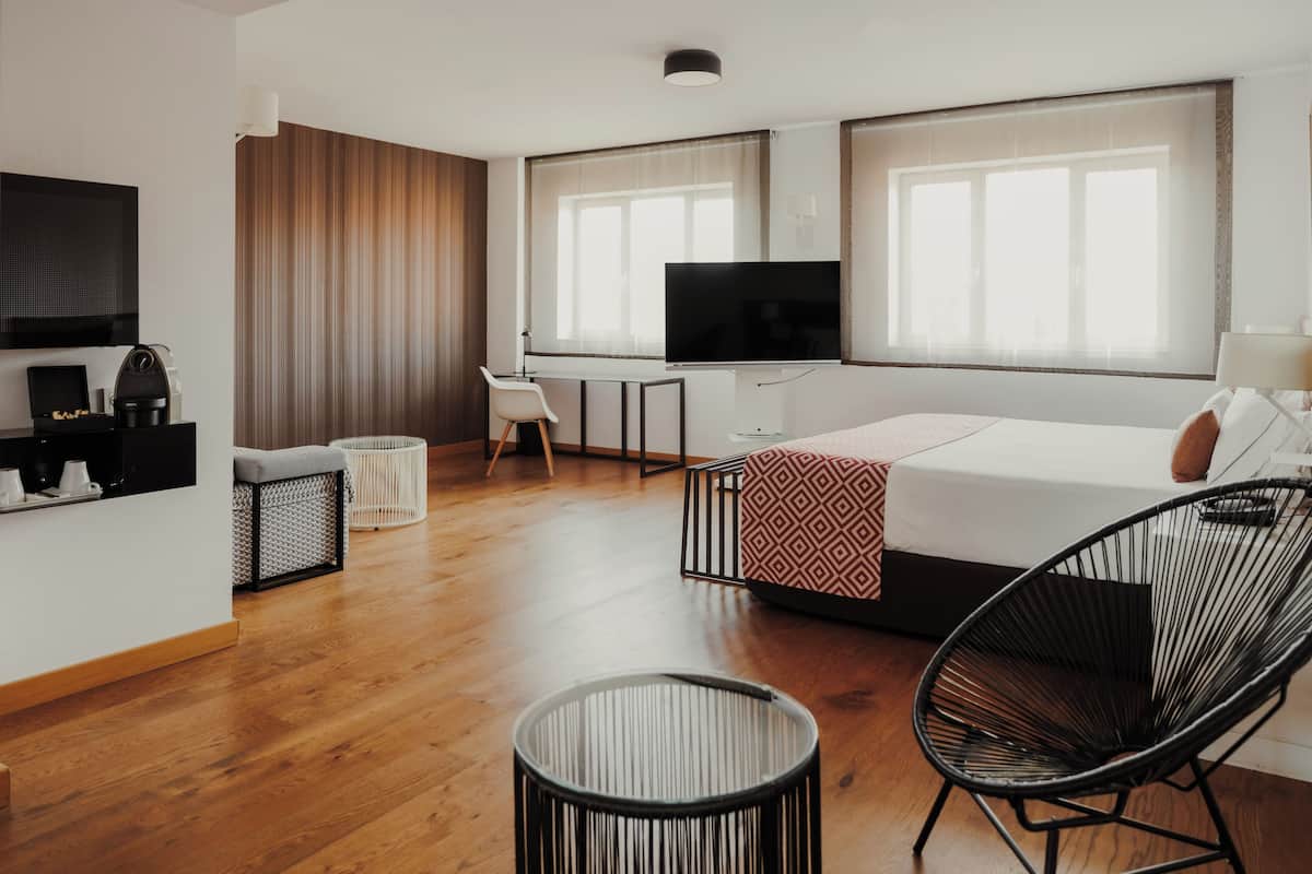A modern hotel room with a wooden floor, a bed with a patterned throw, a black chair, a small table, a TV, and a wall-mounted TV.
