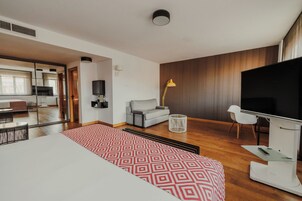A modern hotel room with a large bed, a TV, a sofa, and a dining area with a table and chairs.