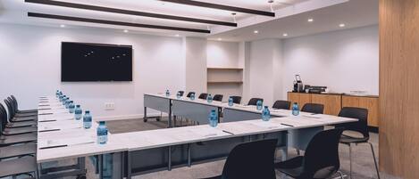 Un salón de conferencias con una larga mesa, sillas, botellas de agua, un televisor de pantalla plana y un armario de madera.