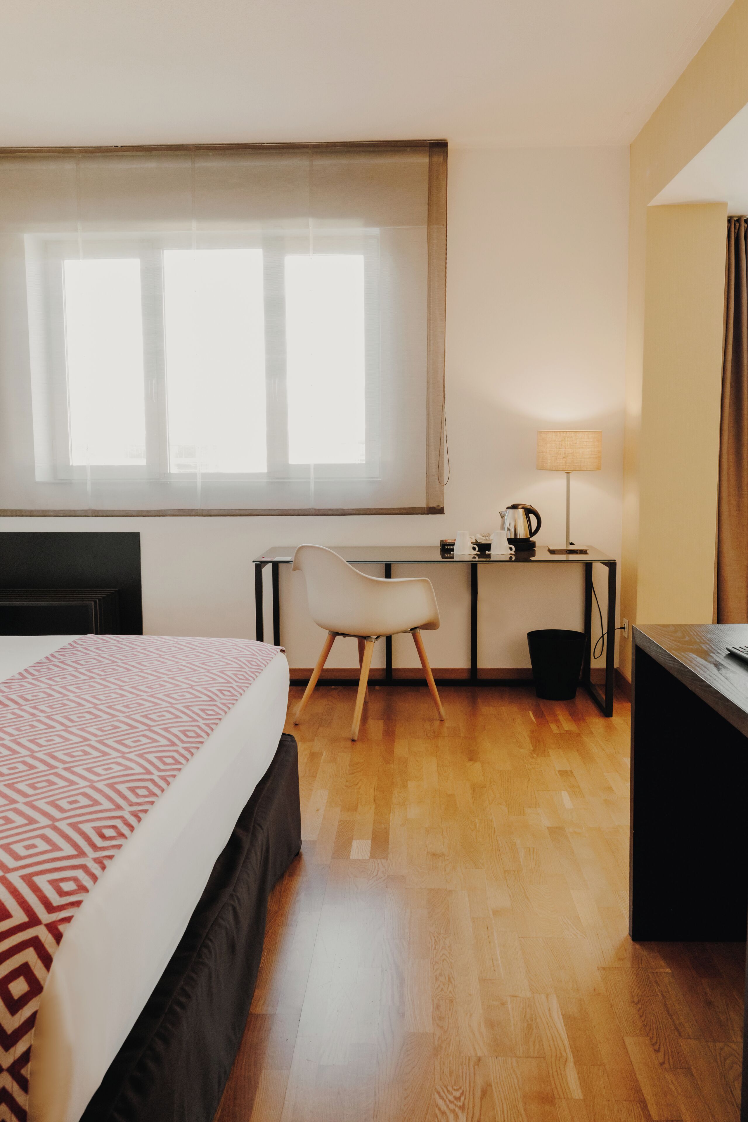 A hotel room with a bed, a desk, a chair, a lamp, and a window with curtains.