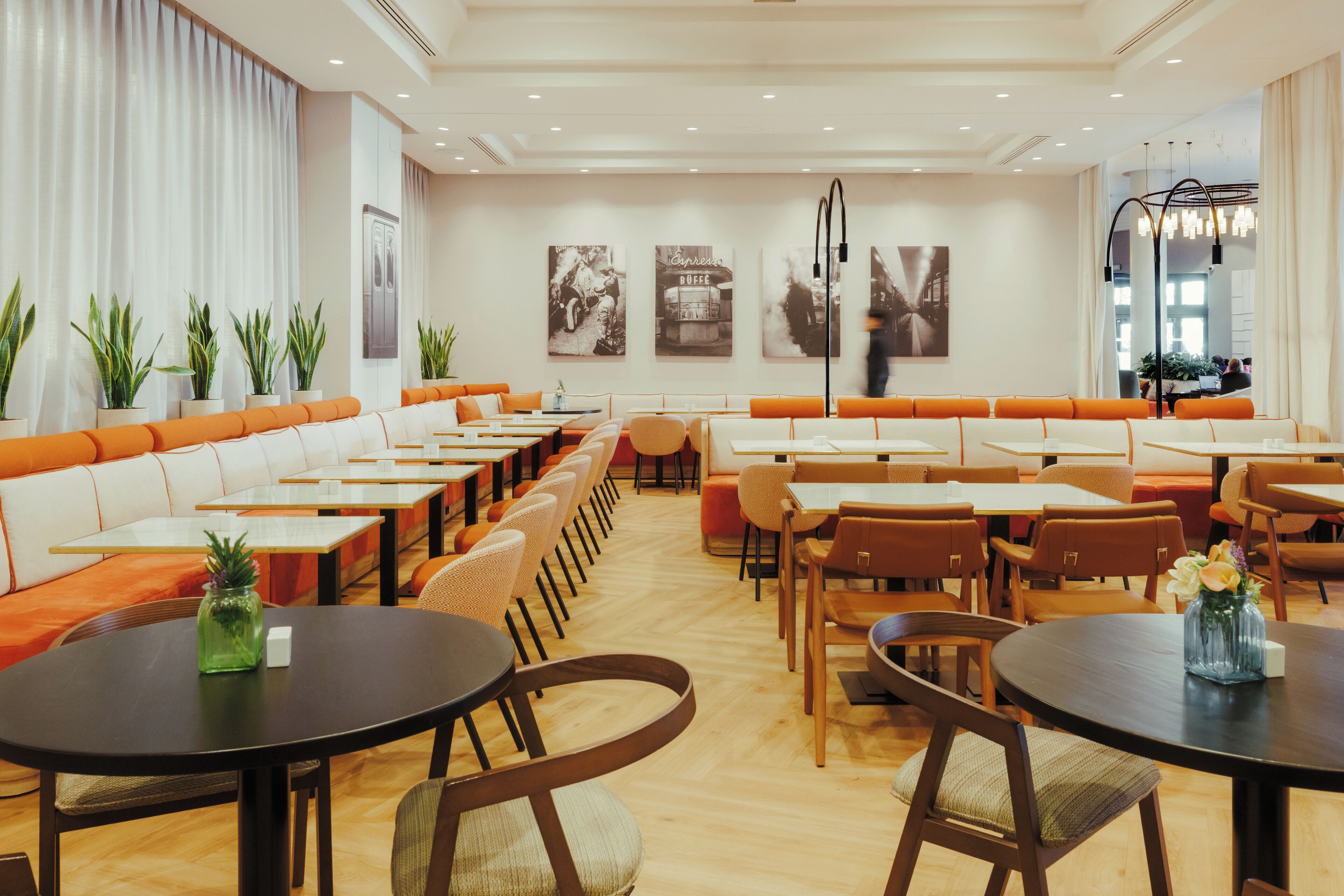 Un restaurante moderno con mesas comunitarias largas y asientos tipo banca color naranja, con un interior limpio y luminoso, suelo de madera y plantas en macetas.