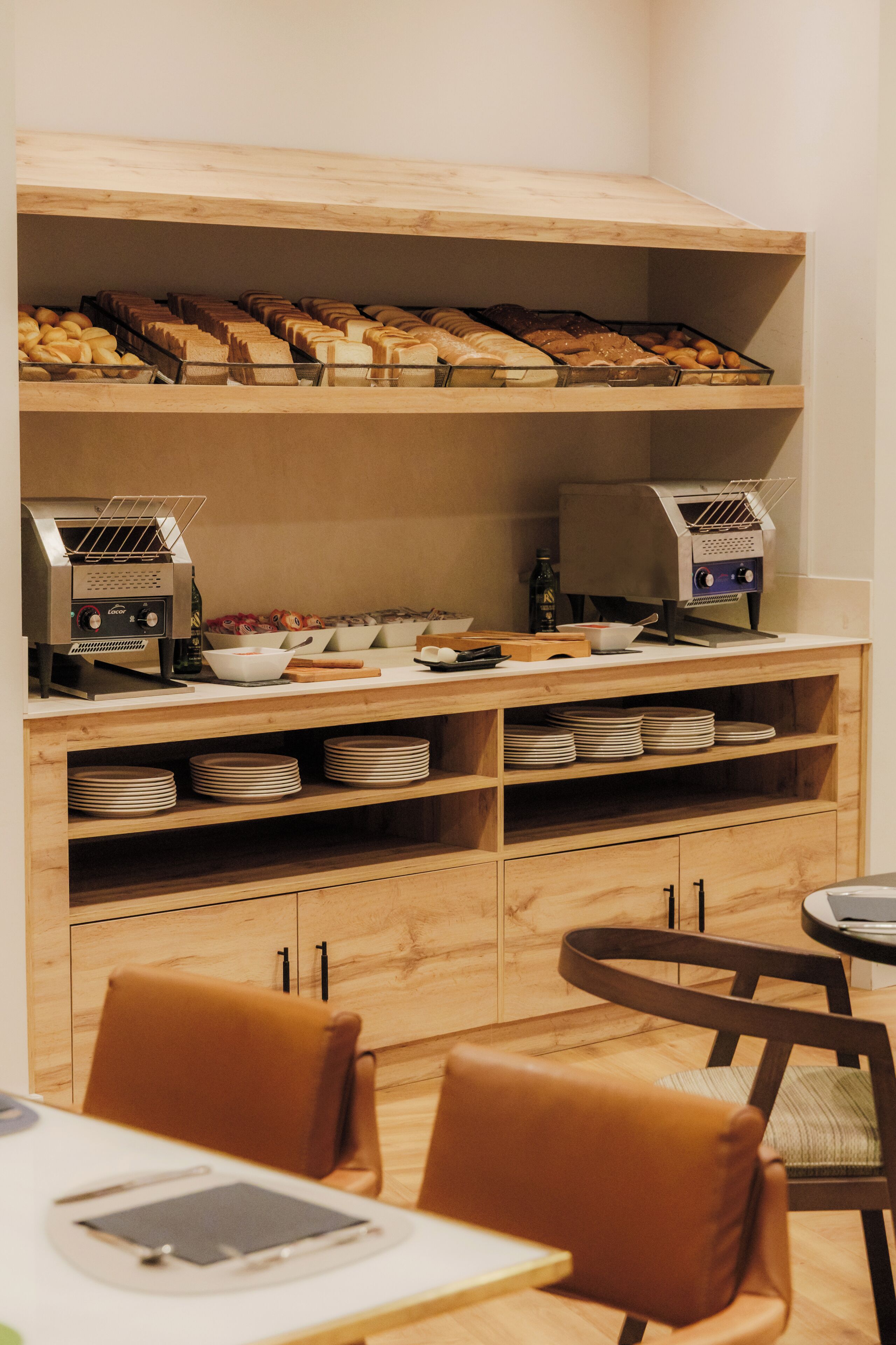 A breakfast buffet area with a variety of breads, pastries, and a coffee machine. There are wooden shelves, a coffee station, and a seating area with chairs.