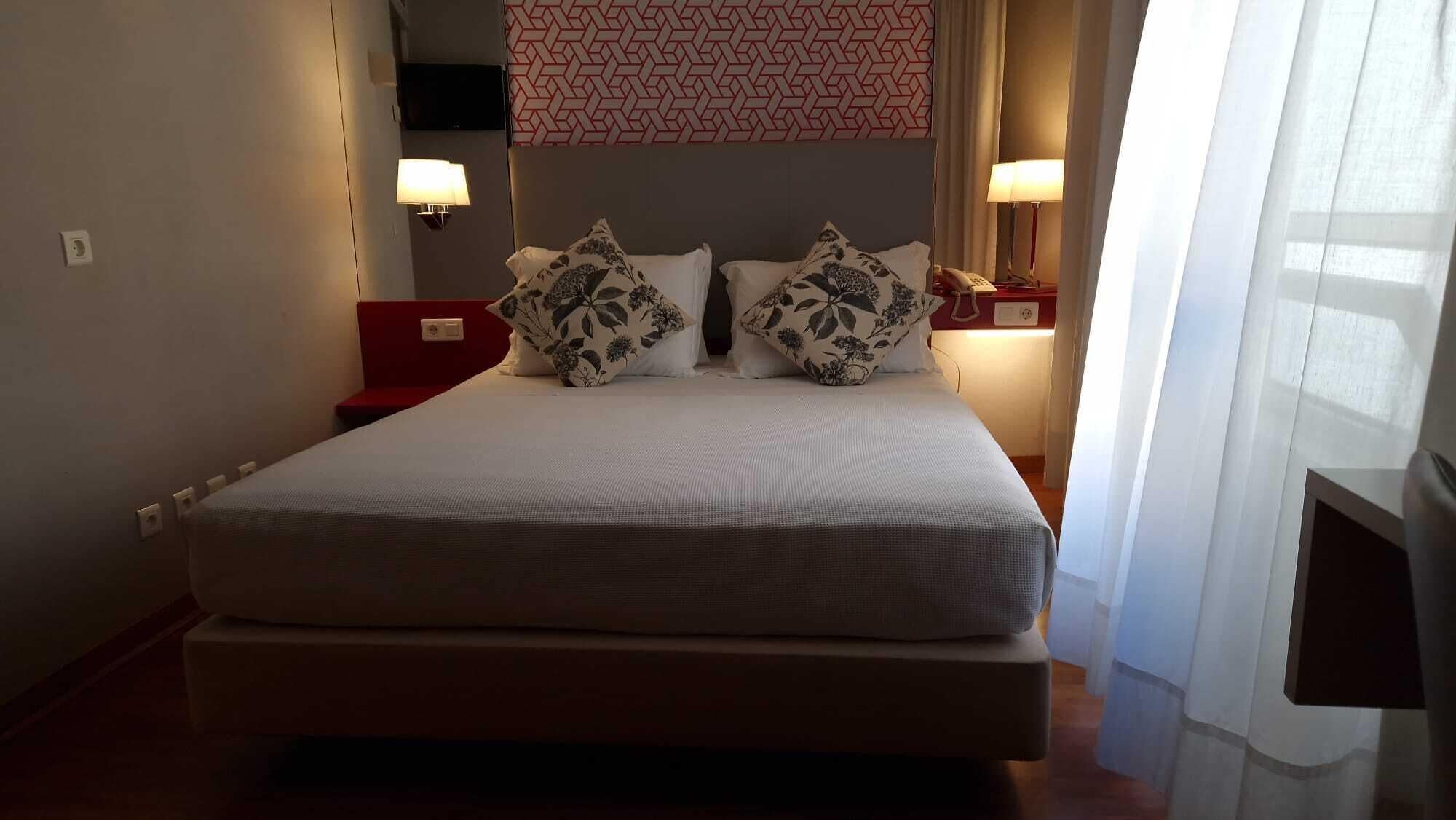 En pænt redt seng med blomsterpuder, et vægmonteret TV og sengeborde med lamper i et hotelværelse.