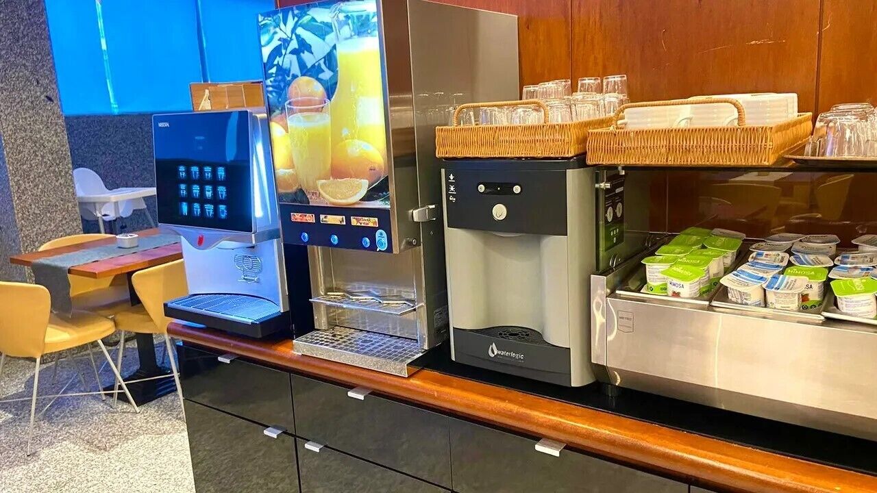 A self-service beverage station with a coffee machine, a juice dispenser, and a refrigerator stocked with yogurt containers.