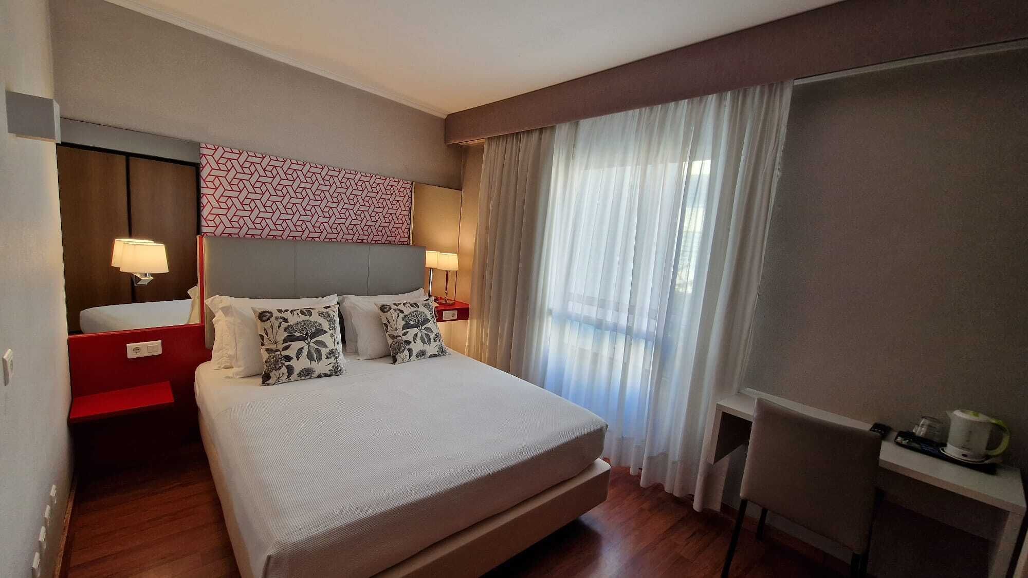 A hotel room with a large bed, two bedside tables with lamps, a red nightstand, a small table with a kettle, and a window with sheer curtains.