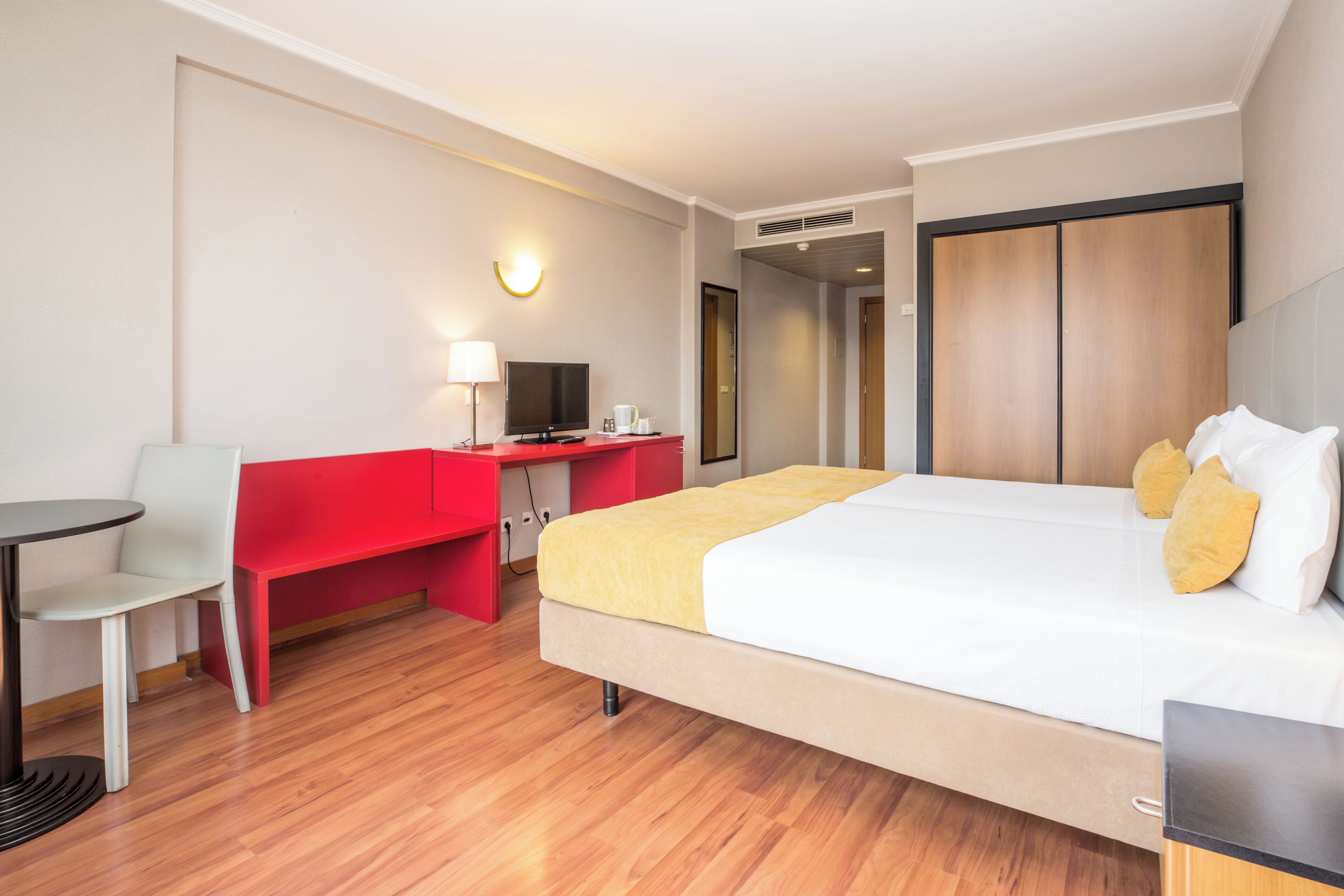 Quarto de hotel com cama, escrivaninha, cadeira e televisão.
