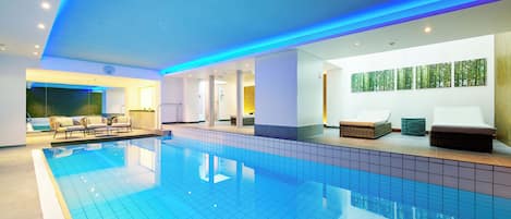 Una piscina interior moderna con suelo de baldosas, sillones de descanso e iluminación azul en el techo.