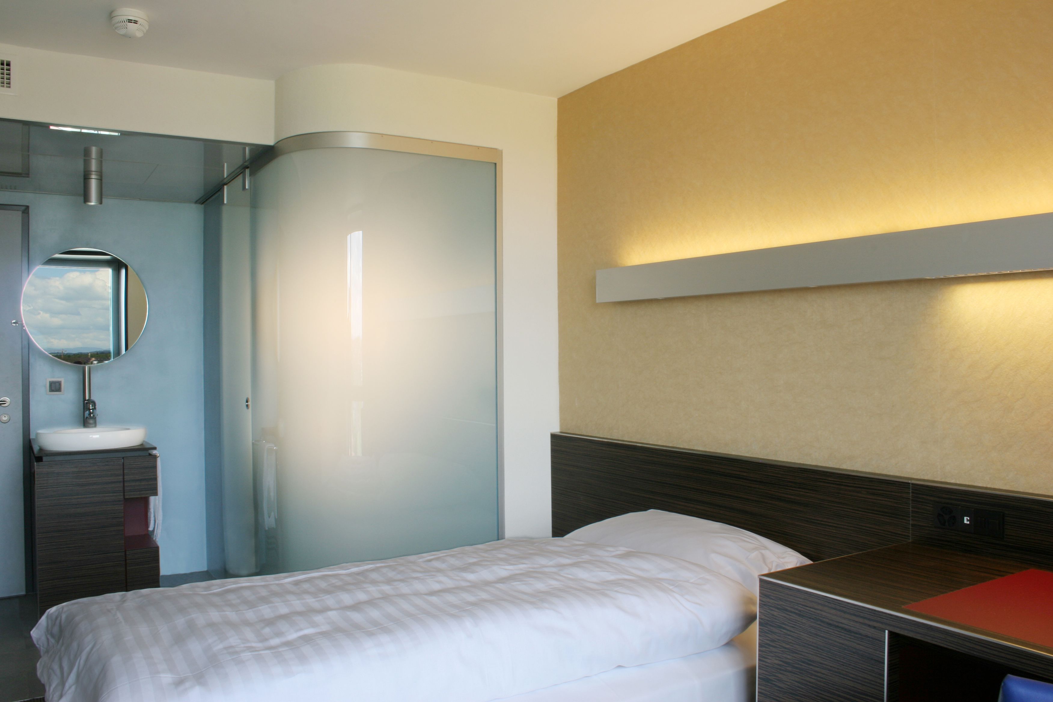 A hotel room with a bed, a bedside table, a mirror, and a bathroom with a sink and glass shower enclosure.